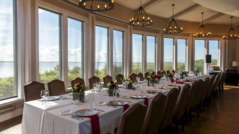 The image shows a dining area set up for a formal event with a long table, chairs, place settings, and large windows offering a scenic view.