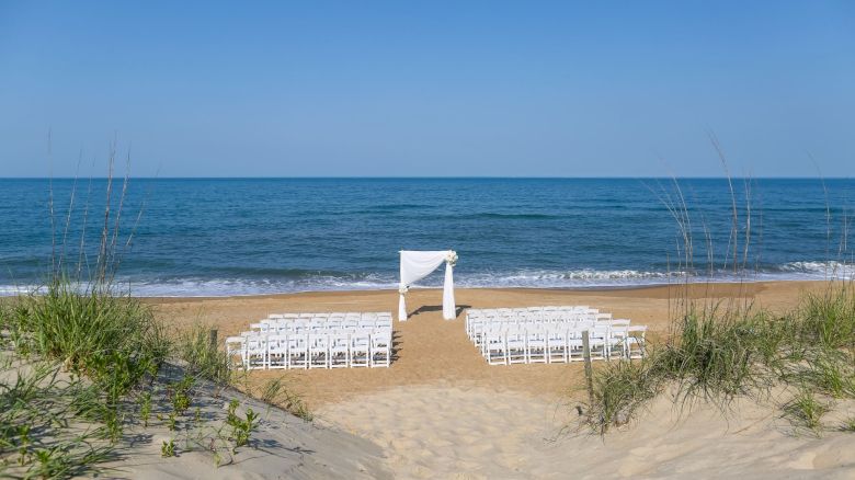 A beach wedding setup with rows of white chairs facing a decorated arch by the ocean shoreline, bordered by sand dunes and greenery.