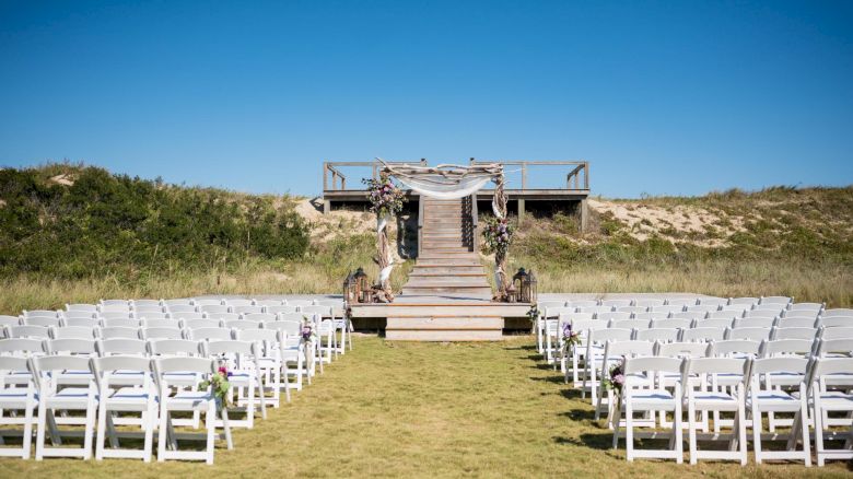 An outdoor wedding setup with rows of white chairs facing a decorated wooden staircase leading to a platform under a clear blue sky.