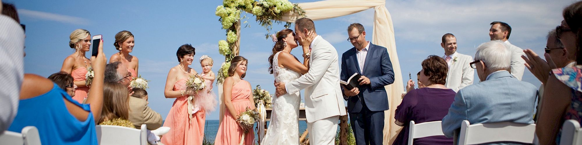 A bride and groom share a kiss at an outdoor wedding ceremony, surrounded by bridesmaids in peach dresses and guests seated on white chairs.