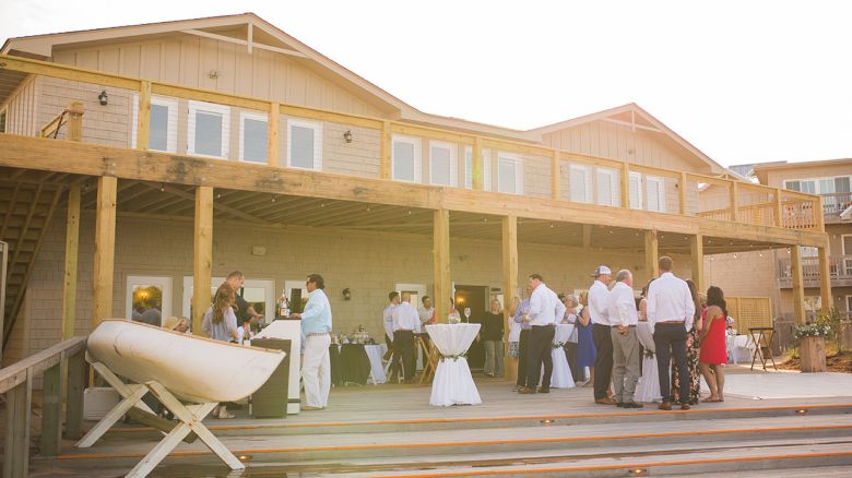 People gather for an outdoor event on a wooden deck in front of a large building with a covered porch and multiple windows.