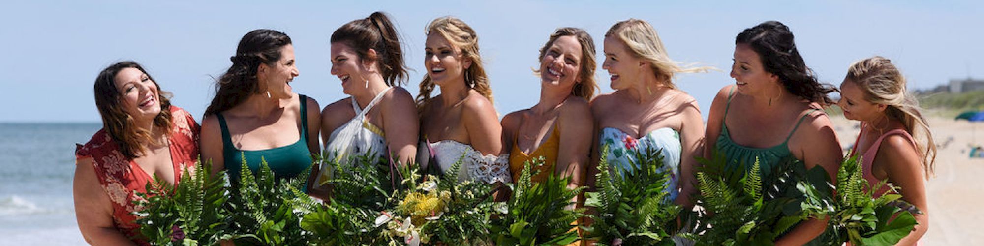 A group of women in colorful dresses holding bouquets, standing together on a beach with the ocean in the background.