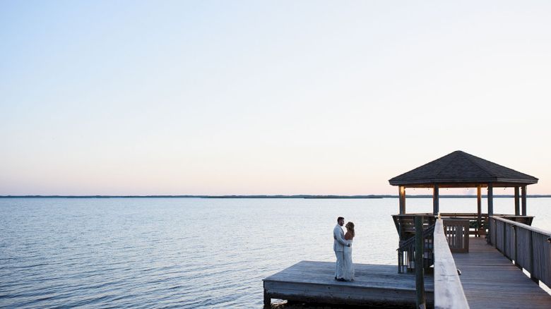 A couple stands embracing on a dock by the water with a gazebo and clear sky in the background.