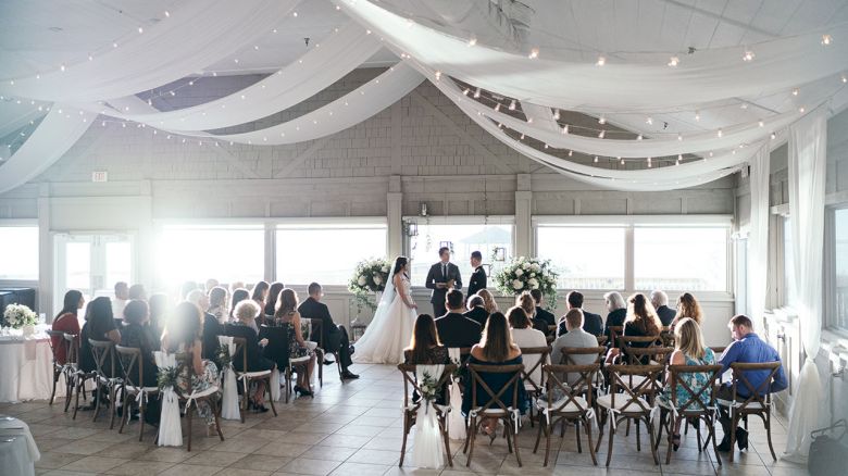 A wedding ceremony is taking place inside a venue with white drapes and string lights, featuring seated guests watching the couple at the altar.