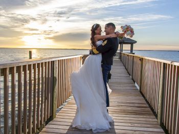 A couple in wedding attire embrace on a wooden boardwalk by the water during sunset, with the bride holding a bouquet.