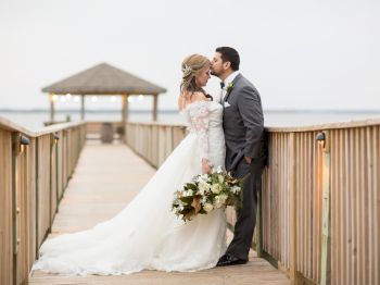 A bride and groom share a kiss on a wooden boardwalk, with the bride holding a bouquet and a gazebo visible in the background by the water.