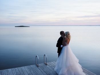 A couple in wedding attire stands on a wooden dock by a serene lake, embracing each other during sunset.