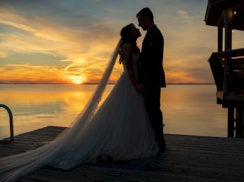 A couple in wedding attire stands on a dock at sunset, sharing an intimate moment by the calm water, creating a romantic silhouette.