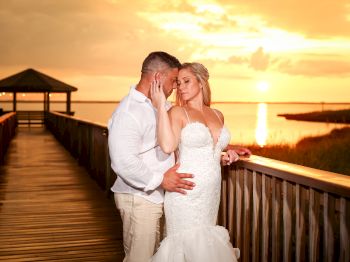 A couple in wedding attire shares an intimate moment on a wooden pier at sunset, with a serene, golden sky in the background.