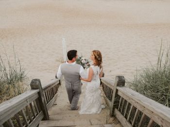 A couple in wedding attire walks down wooden stairs leading to a sandy beach, holding hands. The setting is serene with natural greenery.