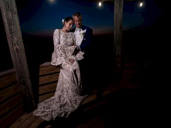 A couple is dressed in formal wedding attire, posing closely together under a wooden structure with dim lighting and a dark background.