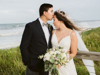 A couple in wedding attire stands by the beach, with the groom kissing the bride’s forehead while she holds a bouquet.
