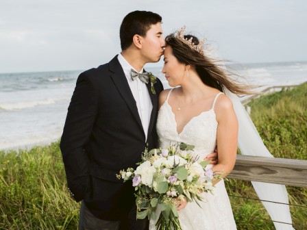 A couple in wedding attire stands by the beach, with the groom kissing the bride’s forehead while she holds a bouquet.