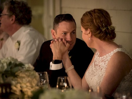 A couple in wedding attire holds hands and gazes at each other lovingly during a reception.