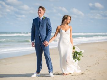 A couple in wedding attire stands on a sandy beach, holding hands with the ocean and blue sky in the background.
