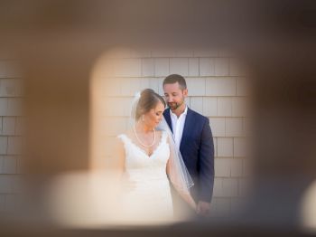 A bride and groom in wedding attire stand close, holding hands, in front of a shingle wall, viewed through an out-of-focus foreground.