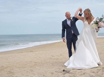 A couple, dressed in wedding attire, is walking on a beach. The bride holds a bouquet of flowers, and they appear happy.
