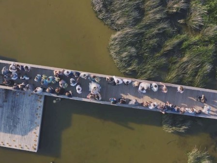 Aerial view of a crowded wooden boardwalk over water, surrounded by vegetation, with some people gathered under a gazebo at the end.