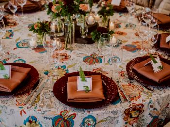 A beautifully set table with floral tablecloth, brown napkins, glassware, small place cards, and green fern leaves at each setting, surrounded by decor.