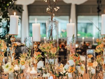 The image depicts a beautifully decorated table with white candles in glass holders and an assortment of flowers in elegant vases.
