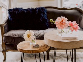 A cozy seating area with a gray couch, dark blue pillows, and round wooden side tables decorated with pink and white flowers in vases.