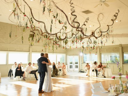 A couple dances at a wedding reception with guests seated around, under a ceiling adorned with hanging decorations and flowers.