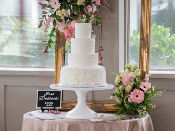 A tiered white cake on a stand is displayed on a table with pink flowers and a 