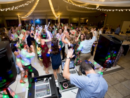 A lively party with a DJ and dancing crowd is taking place under string lights in a decorated indoor venue.