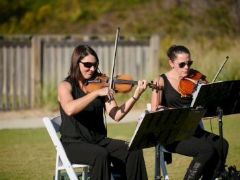 Two violinists are playing music outdoors, both wearing black outfits and sunglasses, with music stands in front of them.