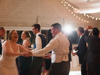 People are dancing joyfully at what appears to be a wedding reception, under string lights in an indoor setting.