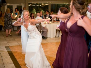 A bride in a white dress dances with two women in purple dresses at a reception, while seated guests watch in the background.