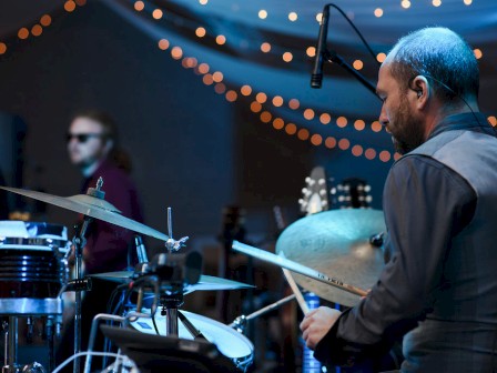 A drummer is playing on stage with other musicians. String lights are hanging in the background, creating a festive atmosphere.