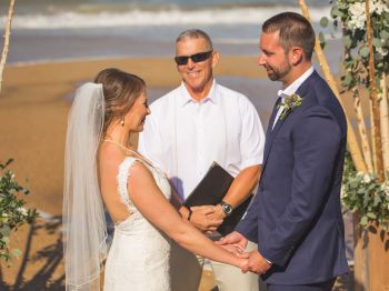 A couple is getting married on a beach with an officiant holding a book, all smiling and holding hands.
