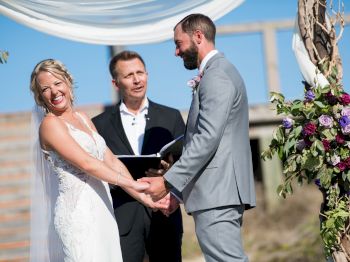 A couple is exchanging vows at an outdoor wedding ceremony, with a pastor officiating. Flower arrangements and a draped canopy decorate the scene.