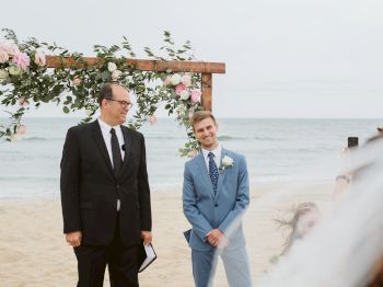 A man in a suit stands next to another man in a blue suit under a floral arch on the beach, with the ocean in the background.
