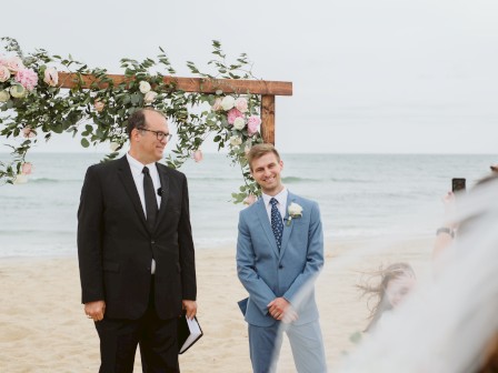 A man in a suit stands next to another man in a blue suit under a floral arch on the beach, with the ocean in the background.