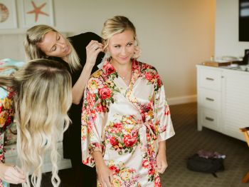 Three women are in a room, two adjusting the hair of the third, who is wearing a floral robe. Items are scattered around.