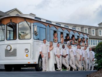 A wedding party stands beside a white trolley bus. Some people are inside the trolley, while others are posed in front of it, all wearing formal attire.