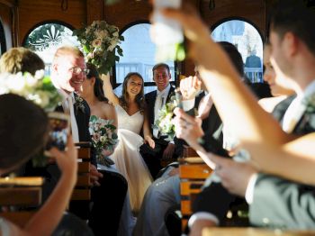 A wedding party rides together in a trolley, with the bride and groom smiling. Guests are holding bouquets and taking photos, celebrating the occasion.