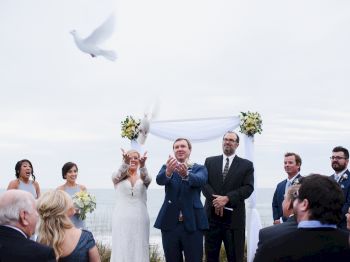 A wedding ceremony with doves being released, surrounded by guests and bridal party near an arch adorned with flowers, outdoors with an ocean view.