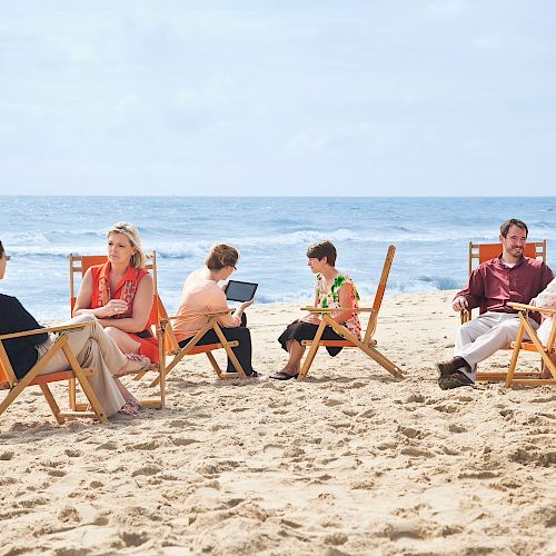 A group of people sit on beach chairs facing each other and engaging in discussions near the ocean on a sunny day.