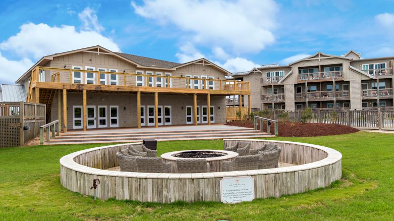 The image shows a large building with balconies, a well-maintained lawn, and a circular seating area around a fire pit in the foreground.
