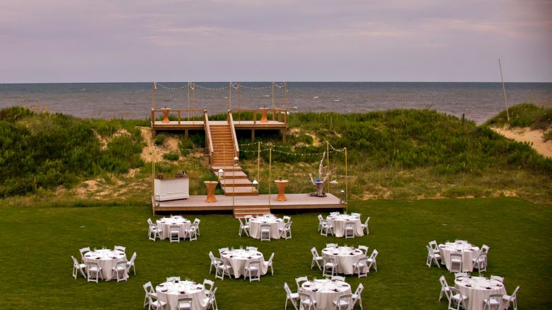 An outdoor event setup on a grassy area by the sea with round tables and white chairs arranged in a semi-circle, and a wooden deck with stairs leading to the beach.