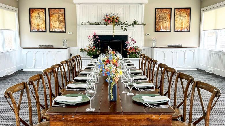 A long dining table is set with plates, napkins, wine glasses, and flowers in a spacious room with high ceilings, fans, and framed artwork on the walls.