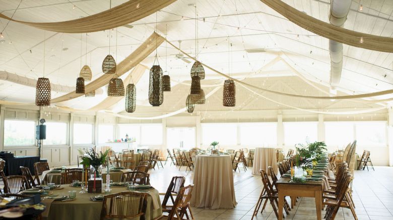 The image shows an elegant event space with draped fabric on the ceiling, lanterns, tables with chairs, and floral centerpieces, creating a festive ambiance.