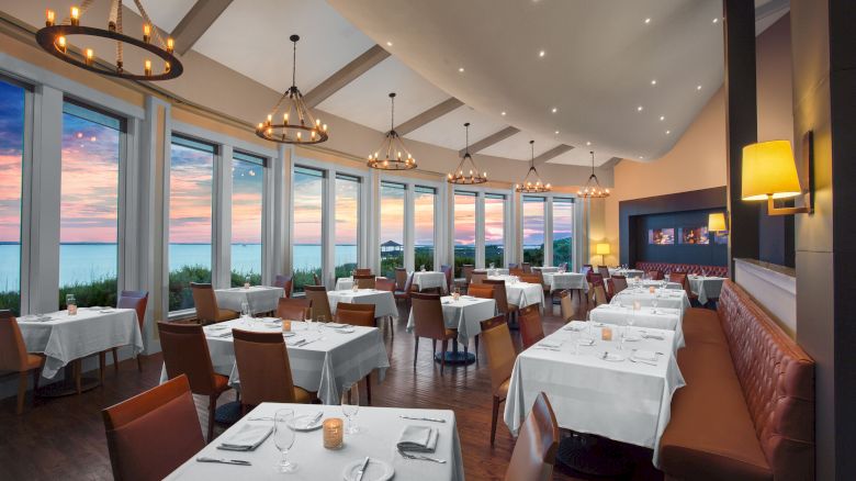 A sophisticated dining area with large windows, set tables, and comfortable chairs, lit with warm lighting and featuring a scenic view of the sunset outside.