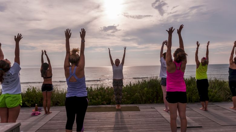 A group of people practicing yoga outdoors near the ocean, stretching their arms upwards with the sun setting in the background, standing in formation.