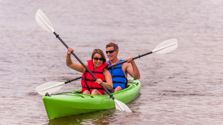 A couple is kayaking together in a green kayak on a body of water, each holding a paddle and wearing life jackets, smiling and enjoying their time.