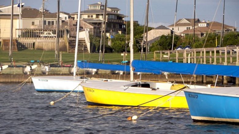 Several colorful sailboats are docked in the water, with houses and greenery visible in the background under a clear sky.