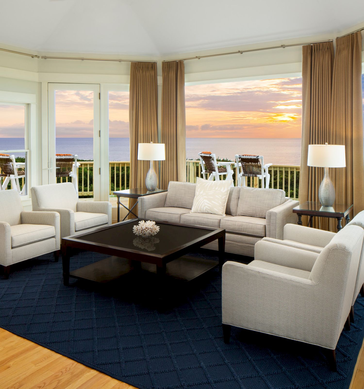 This image shows a modern living room with white furniture, a black coffee table, large windows, and a view of the sunset over the ocean.